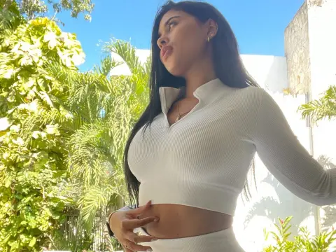 Young woman in a white ribbed long-sleeve crop top posing in a sunny tropical garden setting.