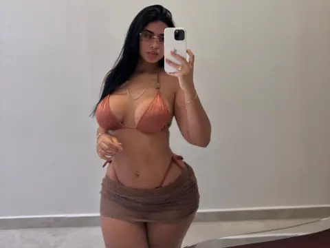 Young woman in a terracotta bikini and crochet mini skirt taking a mirror selfie in a bright room.