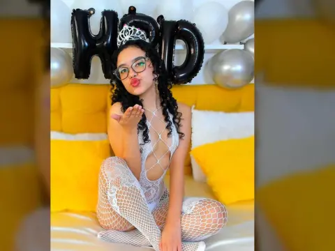 Young woman in white lace bodysuit and fishnet tights sitting on a yellow bed with birthday balloons.