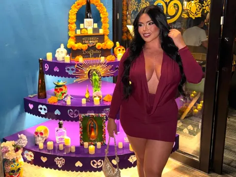 NicoleCruz_ — NicoleCruz_ wears a tight maroon low-cut dress while standing next to a colorful tiered display of skulls and candles.