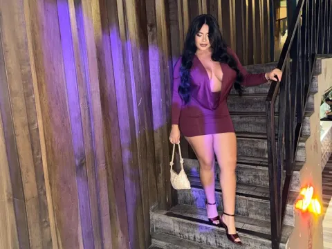 NicoleCruz_ — NicoleCruz_ wears a tight purple dress and high heels while standing on a staircase next to a wooden wall.