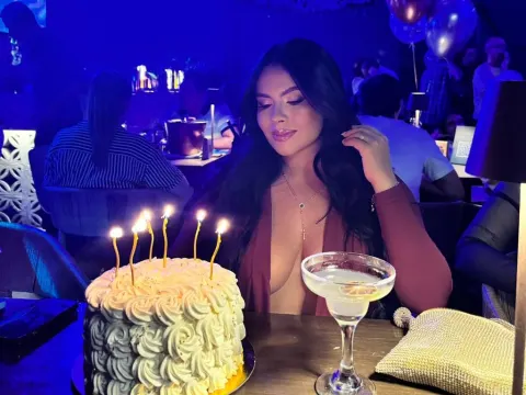 NicoleCruz_ — NicoleCruz_ sits at a table in a low-cut maroon top looking at a lit birthday cake.