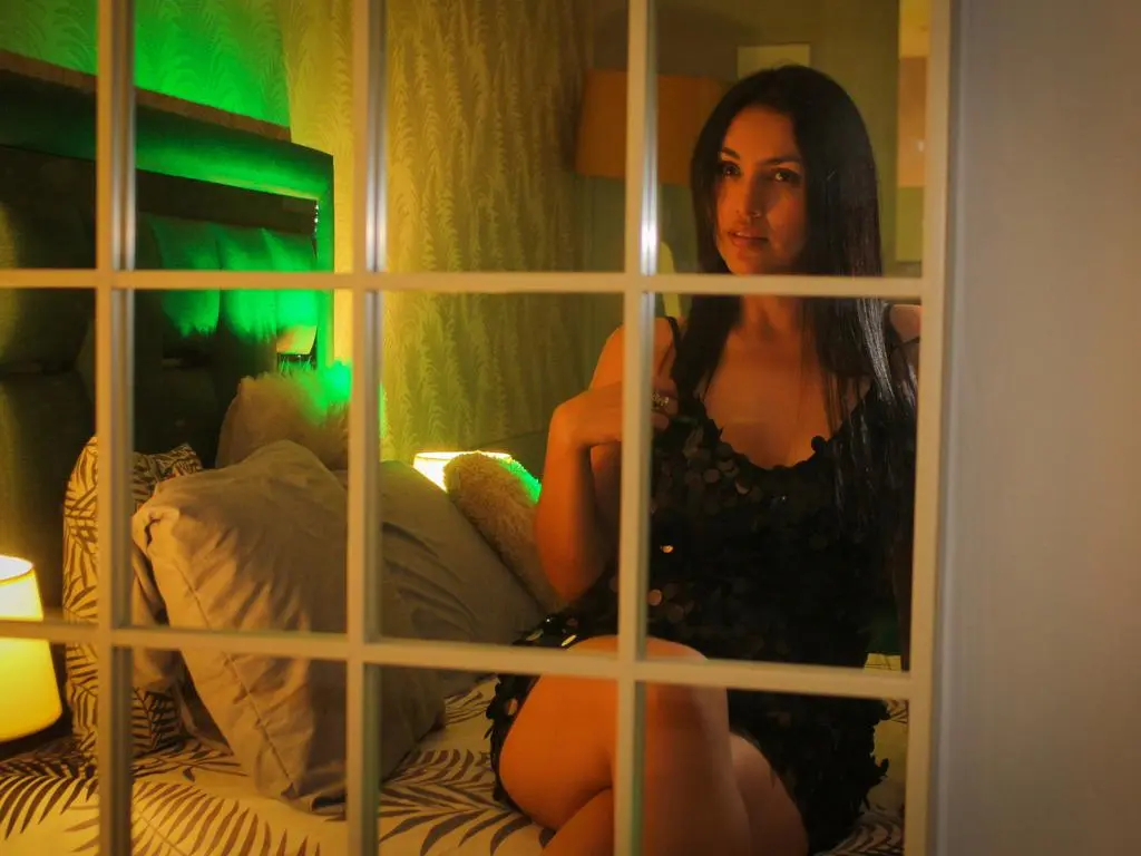 Young woman in a black sequin dress sitting on a bed in a moody, green-lit bedroom.
