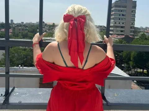 Blonde woman in a red off-the-shoulder dress with a large red bow looking over a city skyline from a rooftop.