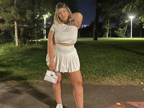 Blonde woman in a white crop top and pleated mini skirt posing in a park at night.