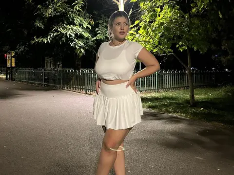 Young woman in a white crop top and mini skirt standing on a night path near trees and a metal fence.