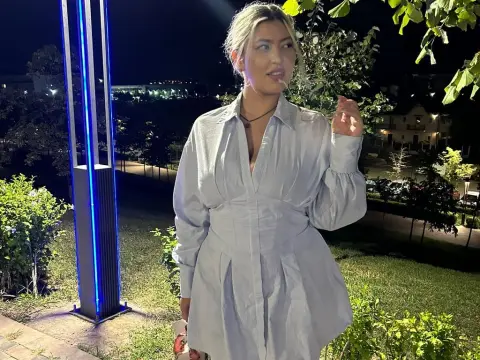 Blonde woman in a white long-sleeve button-down dress standing outdoors at night near blue neon lights.
