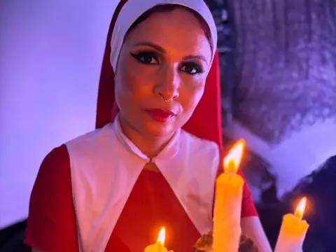 Woman in a red and white nun costume posing by candlelight in a moody, dimly lit setting.