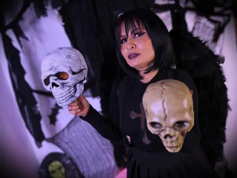 MoiraDevil32 — Woman in black gothic outfit holding a skull prop with dark feathered wings in a spooky Halloween setting.