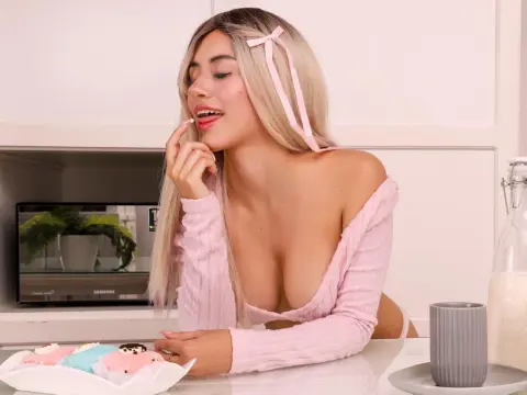 Ari_Love — Blonde woman in a pink off-the-shoulder top eating colorful donuts in a bright, modern kitchen.