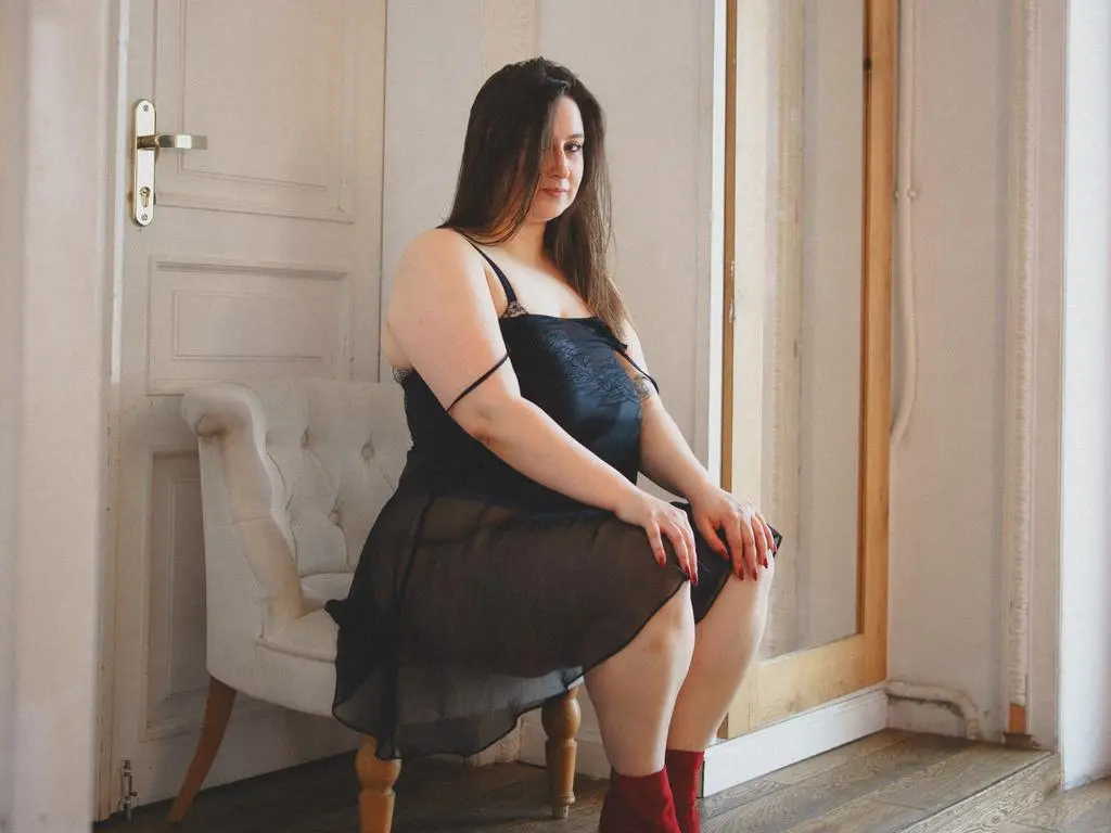 LatteaViia — Curvy woman in a black lace slip dress sitting on a white armchair in a bright, minimalist room.