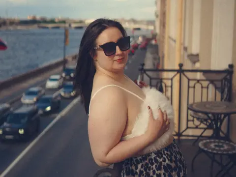 LatteaViia — Smiling woman in sunglasses and a white faux fur top relaxing on a city balcony overlooking a waterfront street.