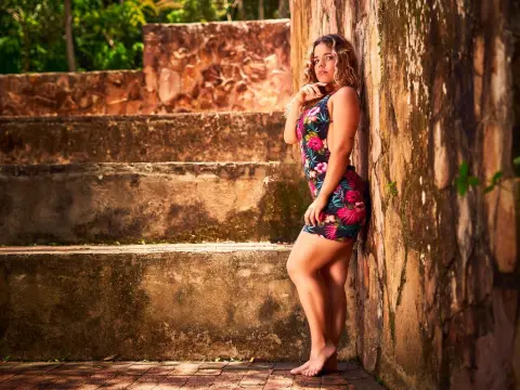 KatalinaCruz — Young woman in a floral summer dress leaning against a rustic stone wall in warm sunlight.