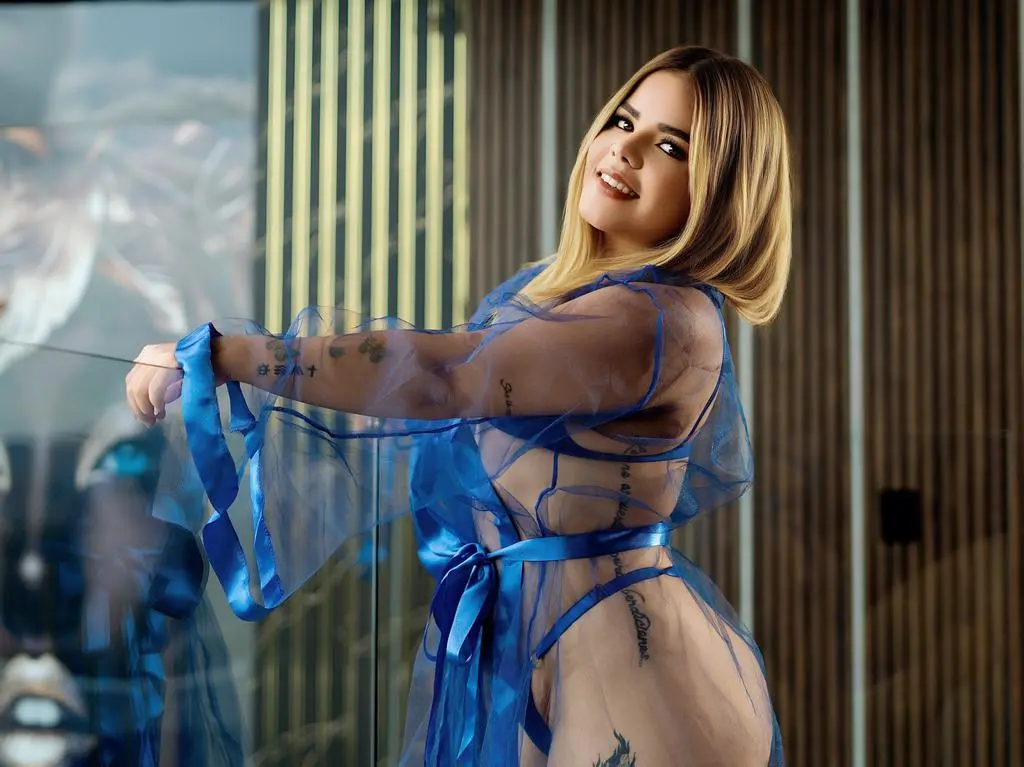 Blonde woman in a sheer blue robe posing elegantly against a modern glass building background.