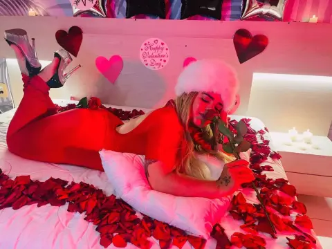 KatalinaCruz — Woman in red outfit lying on rose petals in a romantic pink bedroom for Valentine's Day.