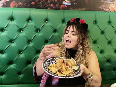 KatalinaCruz — Cheerful woman in a striped top eating a delicious meal in a green tufted booth restaurant setting.