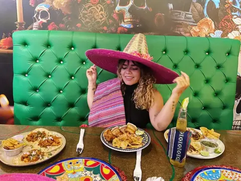 KatalinaCruz — Smiling woman in a wide-brimmed hat enjoying Mexican food and drinks at a vibrant restaurant table.