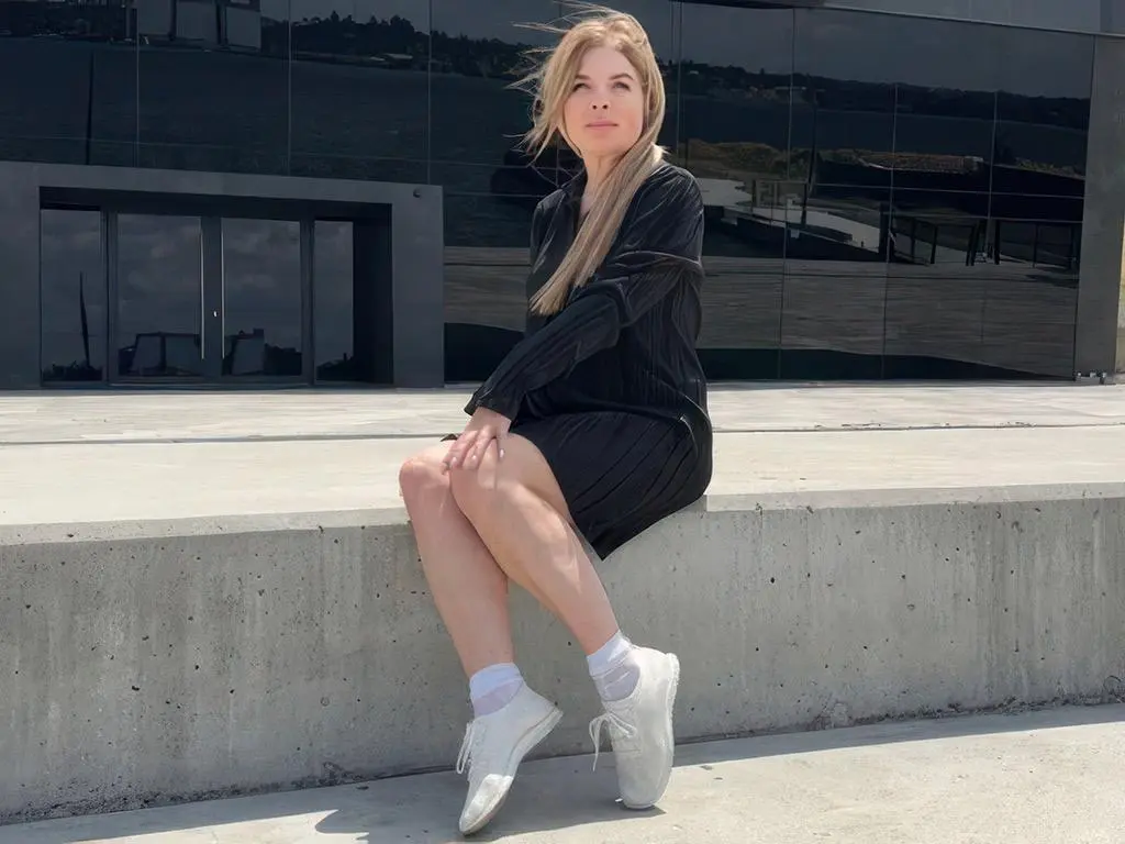 VIKITORY — Blonde woman in a black dress and white sneakers sitting on a concrete ledge by a modern glass building.