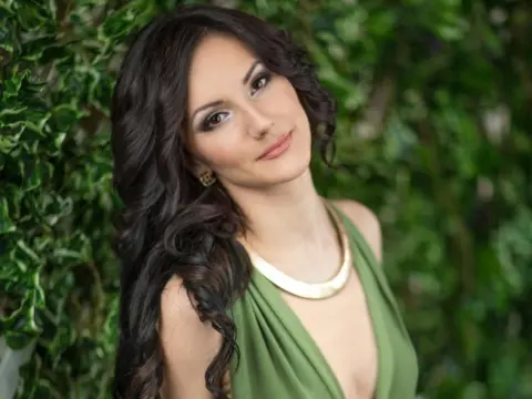 Jaime_Foxy poses with long, wavy dark hair and a gold necklace, wearing a low-cut green top against a leafy green background.