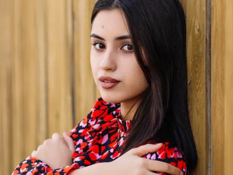 JasmineLove poses with her arms crossed, wearing a red and black patterned top against a wooden fence background.