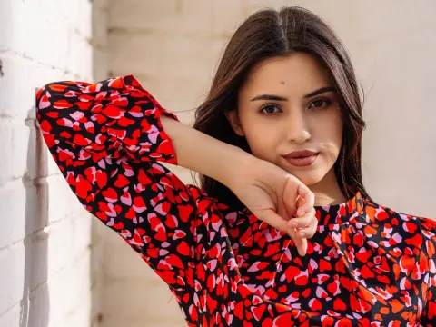 JasmineLove poses outdoors in a red and black patterned blouse with her arm resting against a white brick wall.