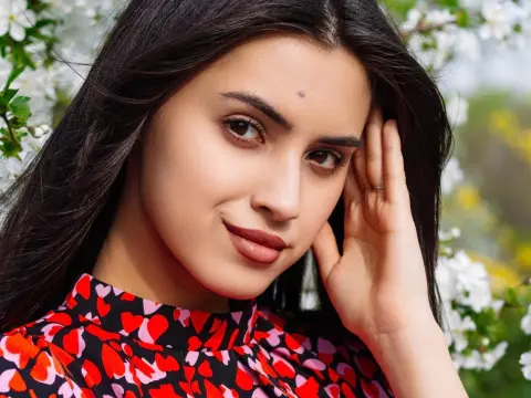 JasmineLove poses outdoors with long dark hair and a red heart-patterned top against a background of white flowers.