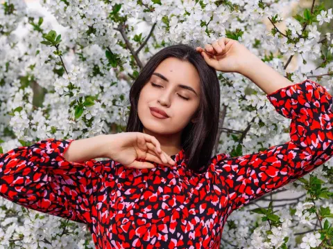JasmineLove poses with her eyes closed in a red and black patterned dress against a background of white flowering trees.
