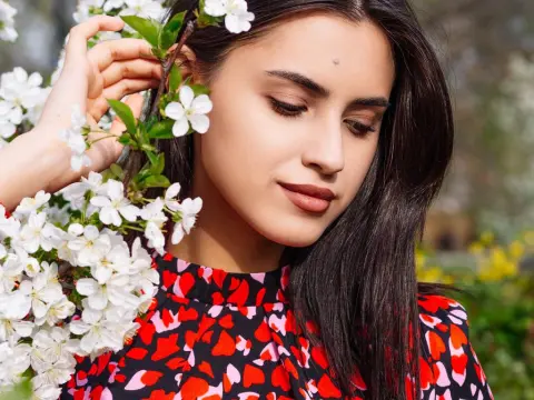 JasmineLove poses outdoors with long dark hair and a red and black patterned top, surrounded by white spring blossoms.