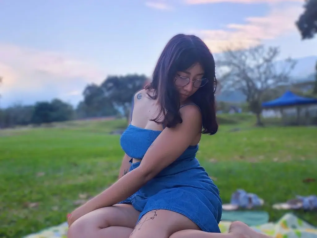 Andromeda_ sits on a blanket in a field wearing a blue denim dress and glasses.