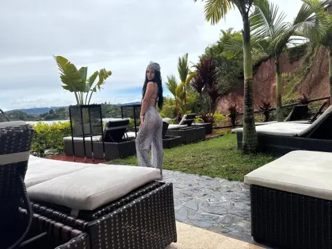 Woman in patterned white trousers standing on a tropical resort patio near palm trees and lounge chairs.