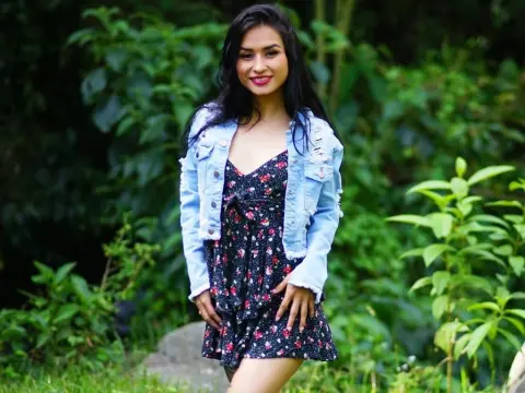 Smiling young woman in a floral sundress and denim jacket standing in a lush green garden setting.