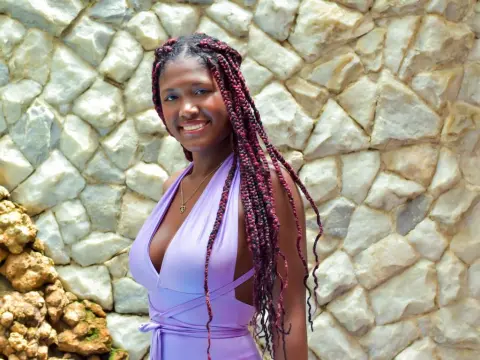 Smiling woman with long burgundy braids wearing a lavender halter dress against a rustic stone wall.