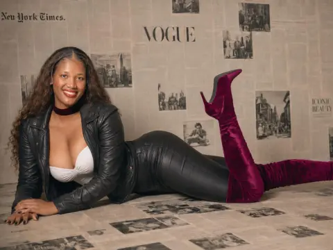 Smiling woman in a black leather jacket and burgundy velvet boots reclining against a Vogue newspaper backdrop.
