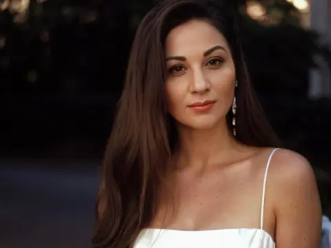 Kethlyne — Elegant woman in a white spaghetti strap dress posing with a serene expression in a soft outdoor setting.