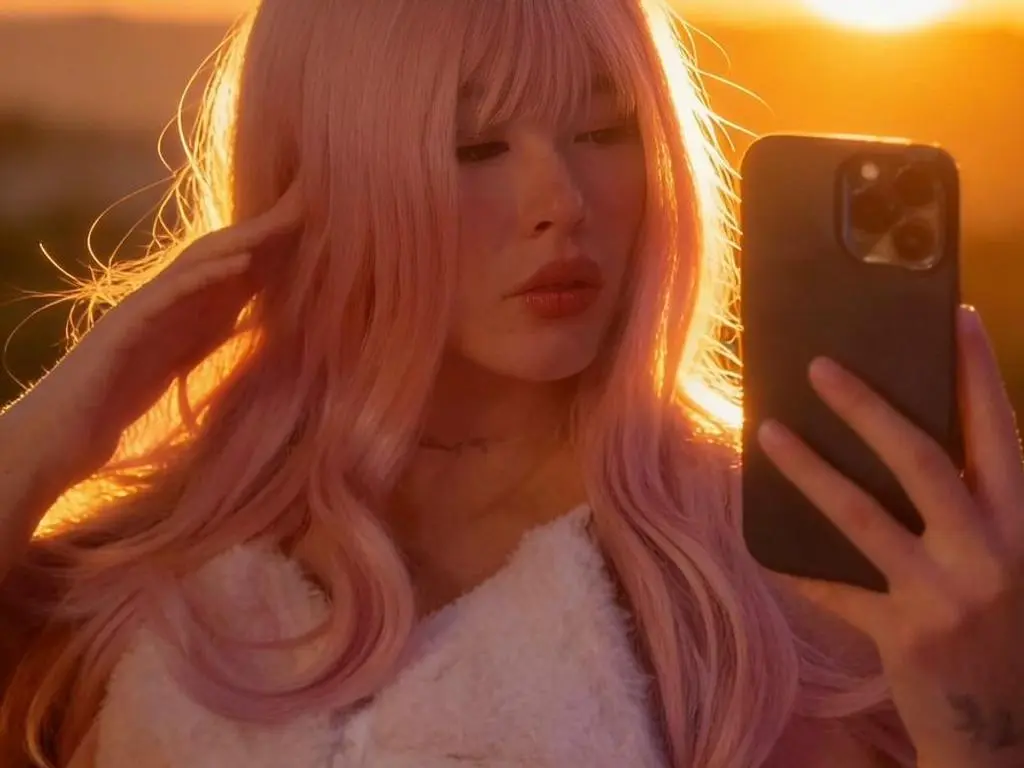 Young woman with long pink hair taking a sunset selfie in a white fuzzy top during golden hour.