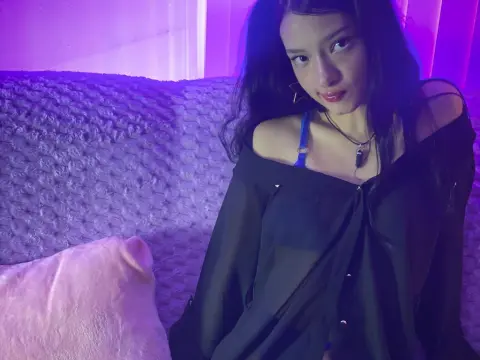 Aurora_Palmer wearing a sheer black button-up shirt and blue bra, sitting on a textured couch under purple lighting.