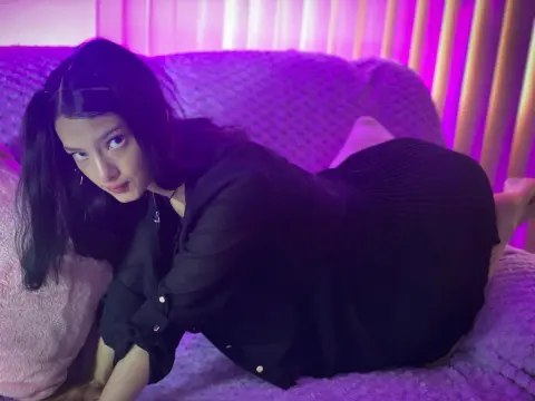 Aurora_Palmer poses on a purple bed wearing a black long-sleeve shirt and black skirt, looking at the camera with long dark hair.