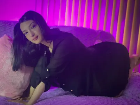 Aurora_Palmer wearing a black long-sleeve shirt while lying on a pink bed against a purple lit background.