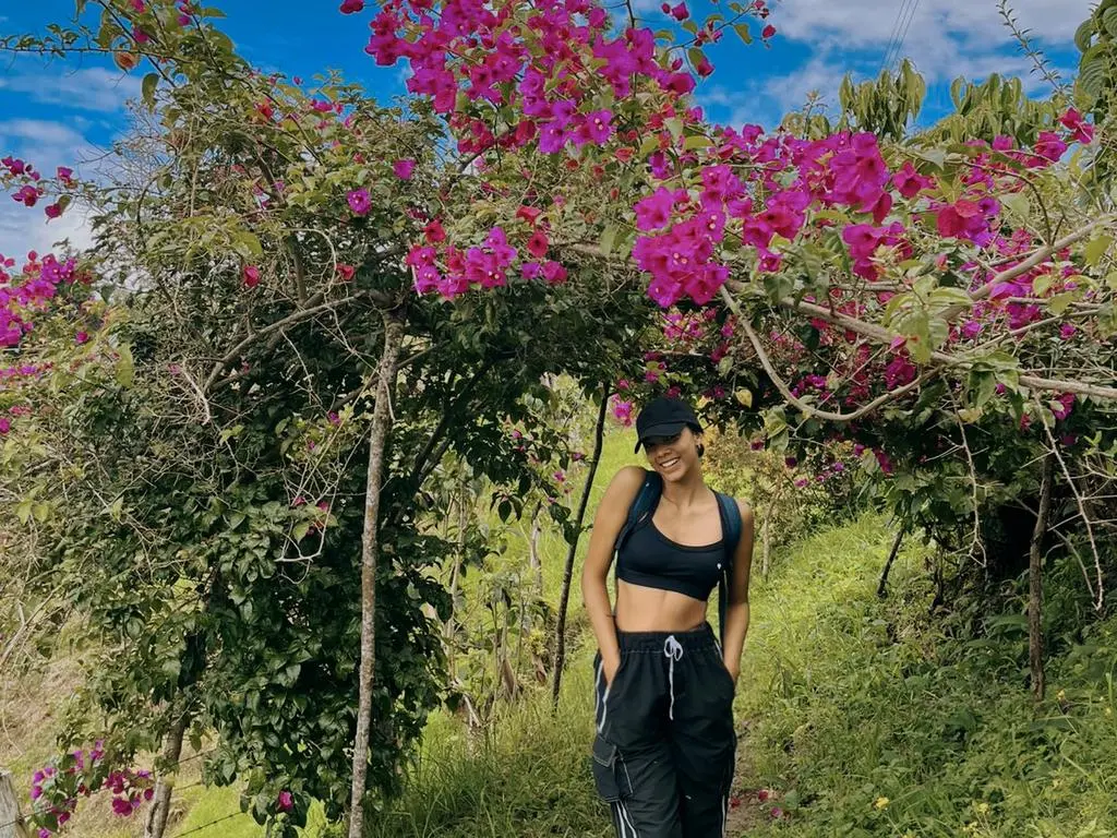 JazzWolstens — Smiling woman in black crop top and cap standing under vibrant pink bougainvillea flowers in a sunny garden.
