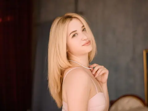 Blonde woman in a pink camisole and pearl necklace posing with a soft, elegant expression in a cozy room.