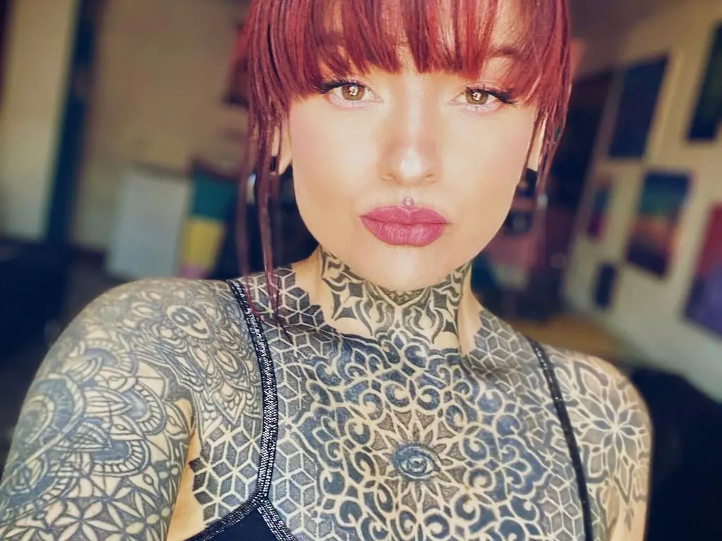 LolaRouse — Close-up portrait of a woman with red hair and intricate black ink tattoos wearing a black tank top.