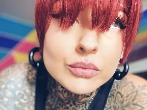 LolaRouse — Close-up of a woman with red hair and facial piercings wearing a black top in a brightly lit setting.