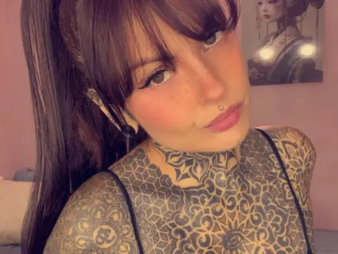 LolaRouse — Woman with long brown hair and extensive neck tattoos wearing a black strap in a cozy indoor setting.