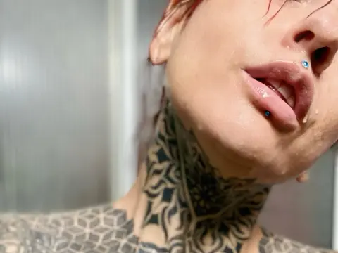 LolaRouse — Close-up of a woman with intricate neck tattoos and a blue nose stud in a bright, minimalist setting.