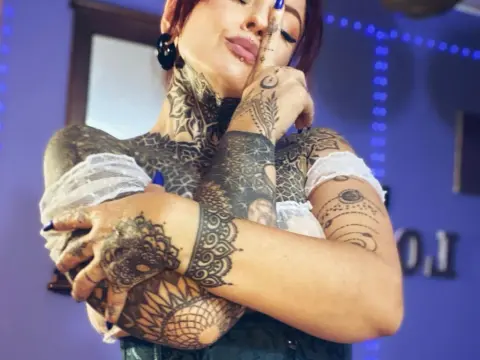 LolaRouse — Tattooed woman in a white top posing with arms crossed against a purple background.