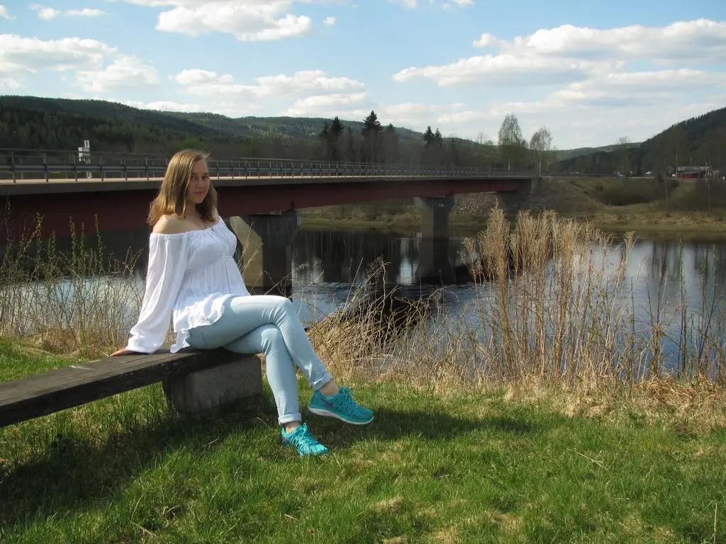 SquirtyAlice — Young woman in white off-the-shoulder top and light jeans sitting on a bench by a river and bridge on a sunny day.