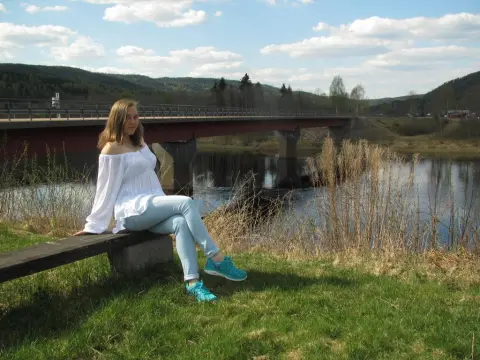 SquirtyAlice — Young woman in white off-the-shoulder top and light jeans sitting on a bench by a river and bridge on a sunny day.