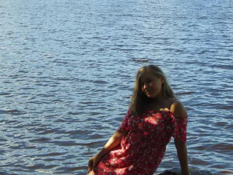 SquirtyAlice — Young woman in a red floral summer dress relaxing by the sparkling blue lake on a sunny day.