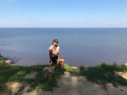 SquirtyAlice — Young woman in casual summer outfit sitting on a rock overlooking the calm blue ocean under a clear sky.