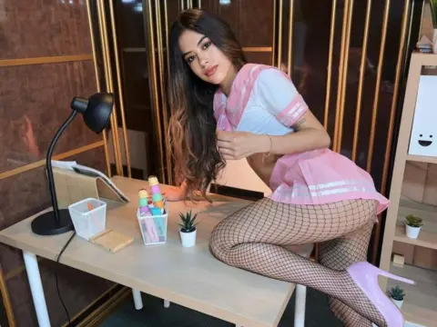SashaCollins — Young woman in a pink schoolgirl outfit and fishnet tights sitting at a desk in a modern office setting.
